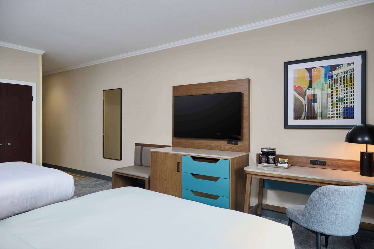 A hotel room with two beds, a desk, a chair, a TV, a coffee maker, and a framed picture on the wall.