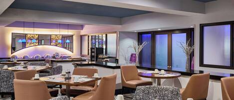 A modern dining area with multiple tables and chairs, a decorative wall with a light fixture, and large windows with blinds.