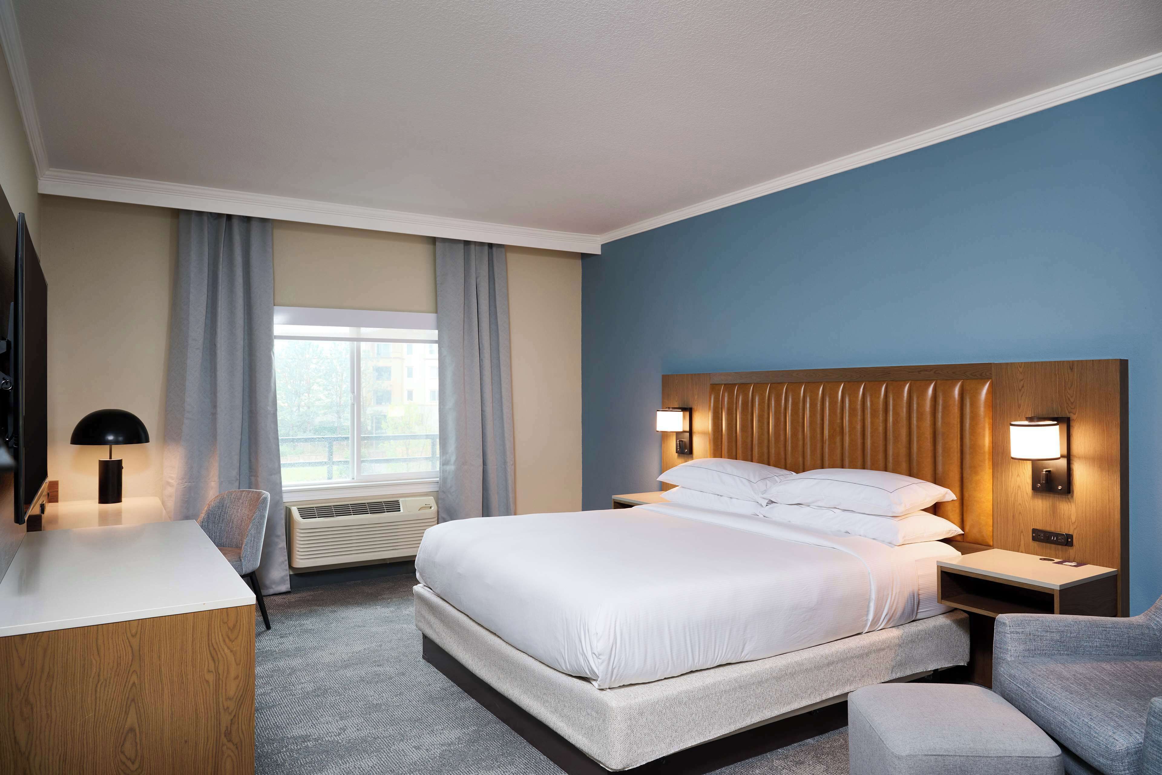 A hotel room with a large bed, a desk, a chair, a nightstand, and a wall-mounted lamp.