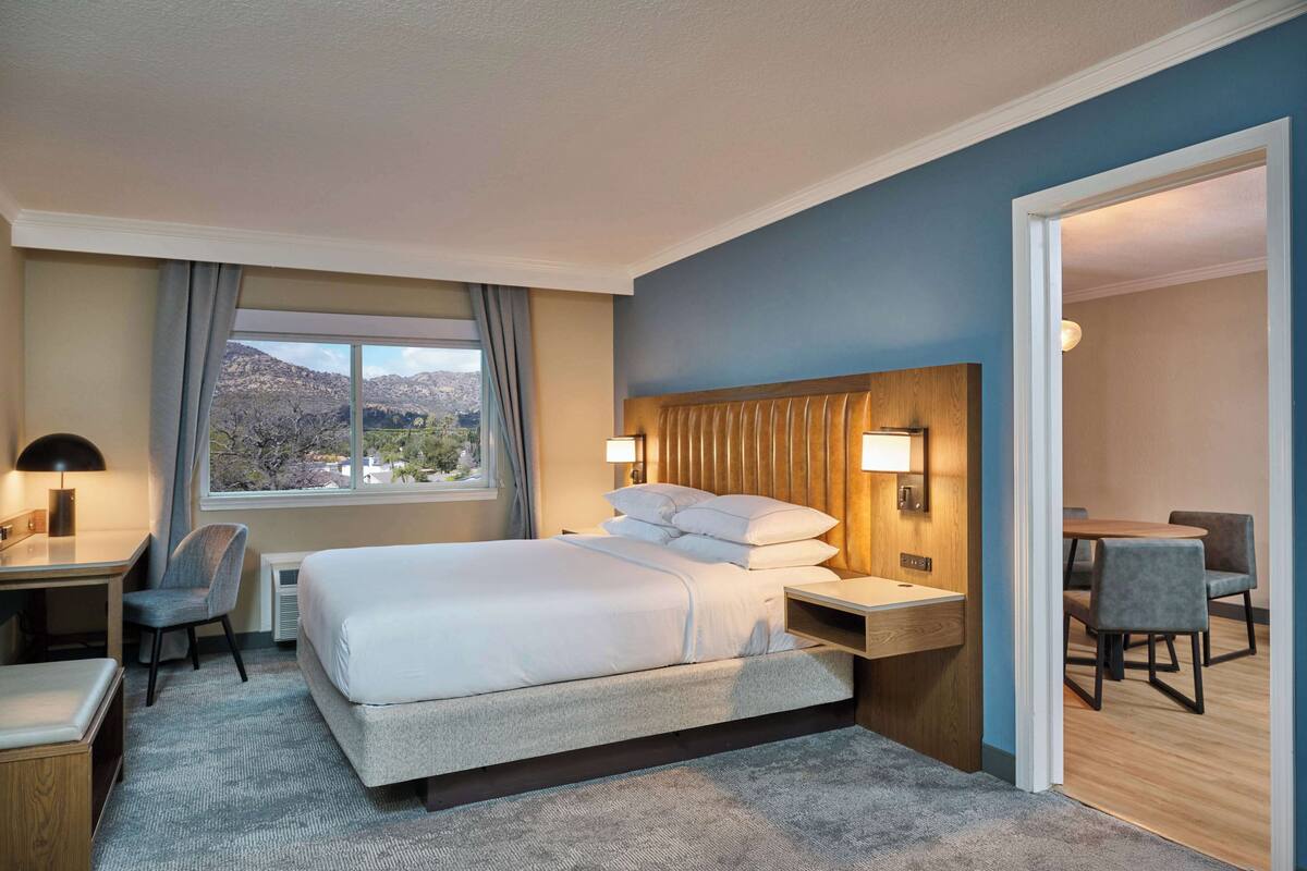 A hotel room with a large bed, a desk, a chair, and a view of mountains through the window.