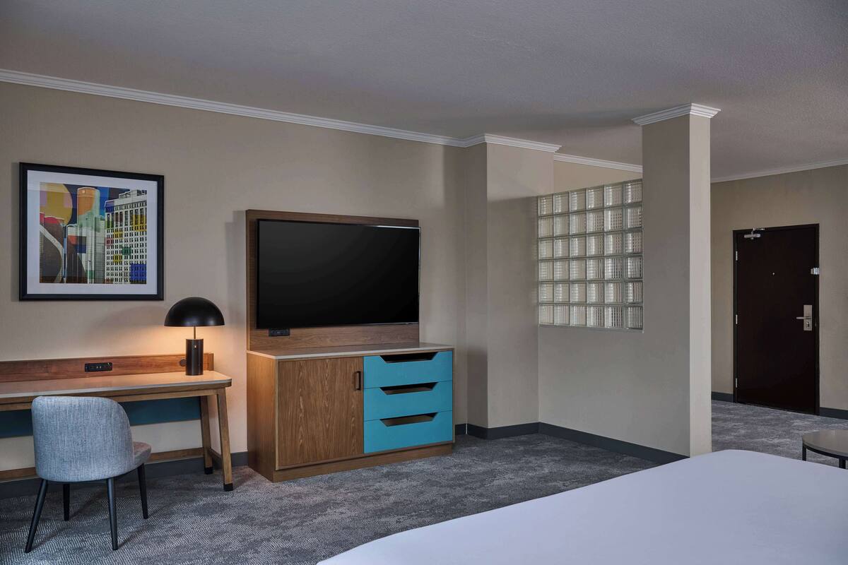 A hotel room with a bed, a desk with a chair, a television, a cabinet with drawers, and a framed picture on the wall.