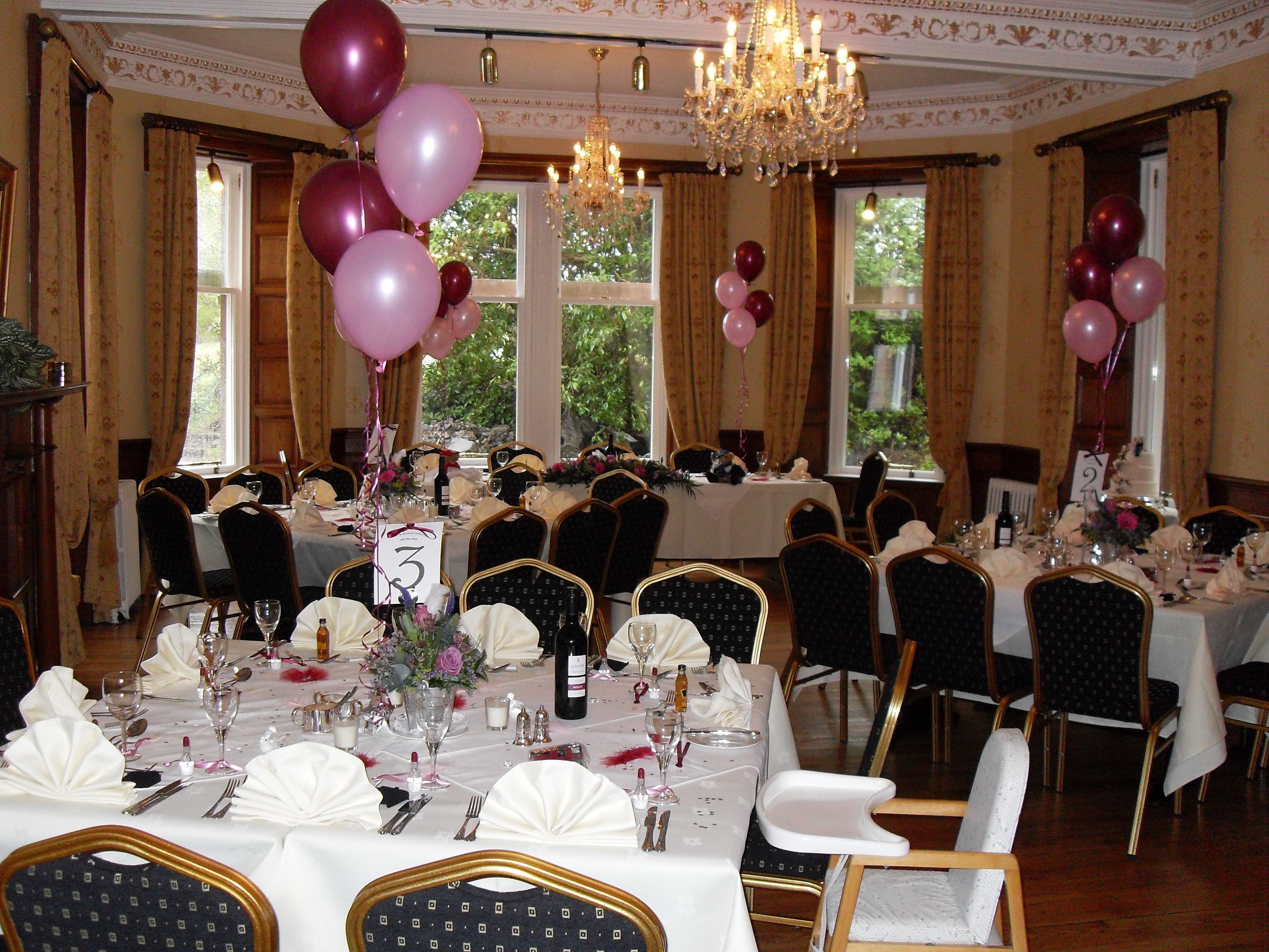 A formal dining room with tables set for a banquet, chandeliers, and large windows with curtains.