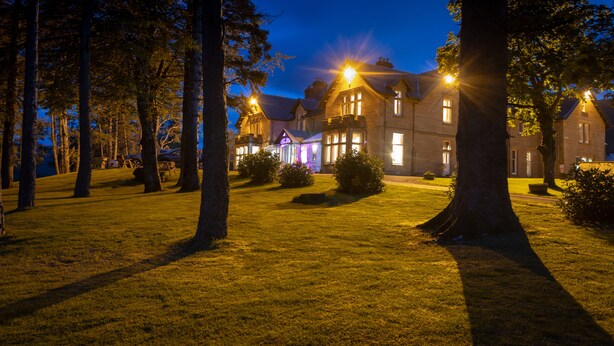 A large, well-lit building with multiple windows and a lawn in the foreground. - Ledgowan Lodge Hotel (Achnasheen)