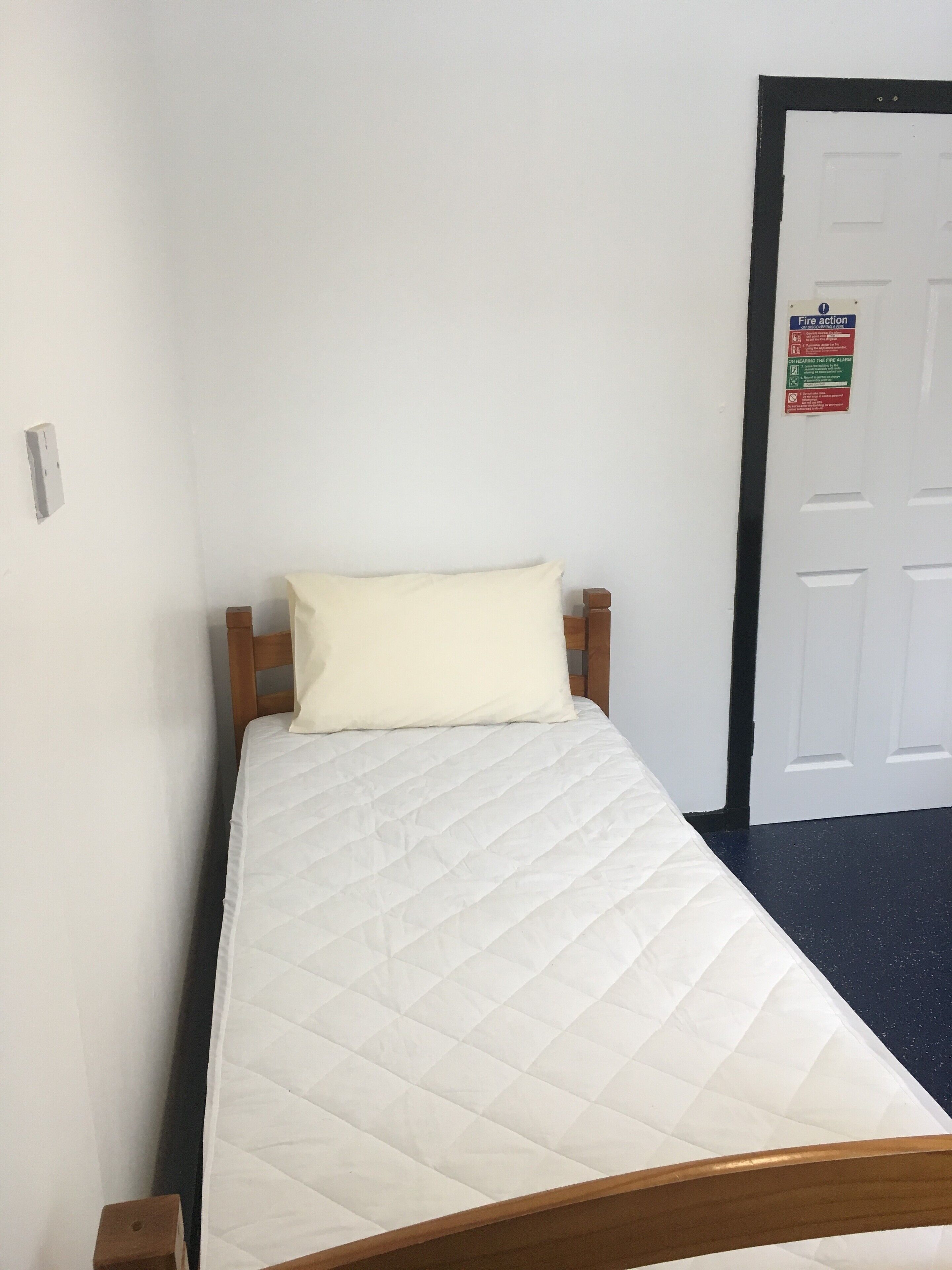 Ein Einzelbett mit gesteppter Matratze und einem Holz-Kopfbrett. Ein weißes Kopfkissen liegt auf dem Bett. Im Hintergrund ist eine Tür mit einem Brandschutzschild zu sehen.