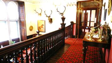 A grand hallway with a red patterned carpet, wooden banister, and mounted animal heads on the wall.