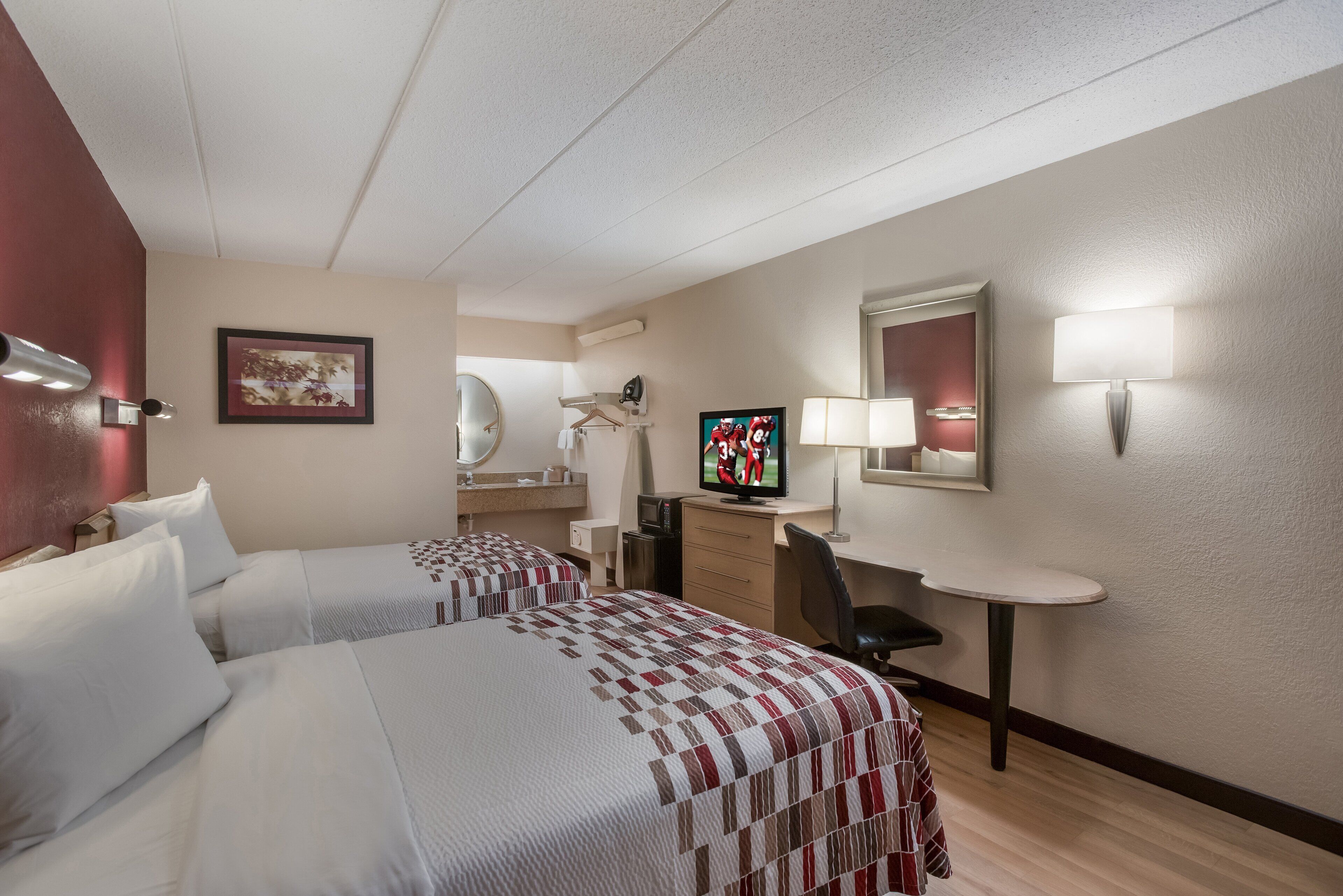Quarto de hotel com duas camas, uma escrivaninha, uma cadeira, uma televisão e um espelho.