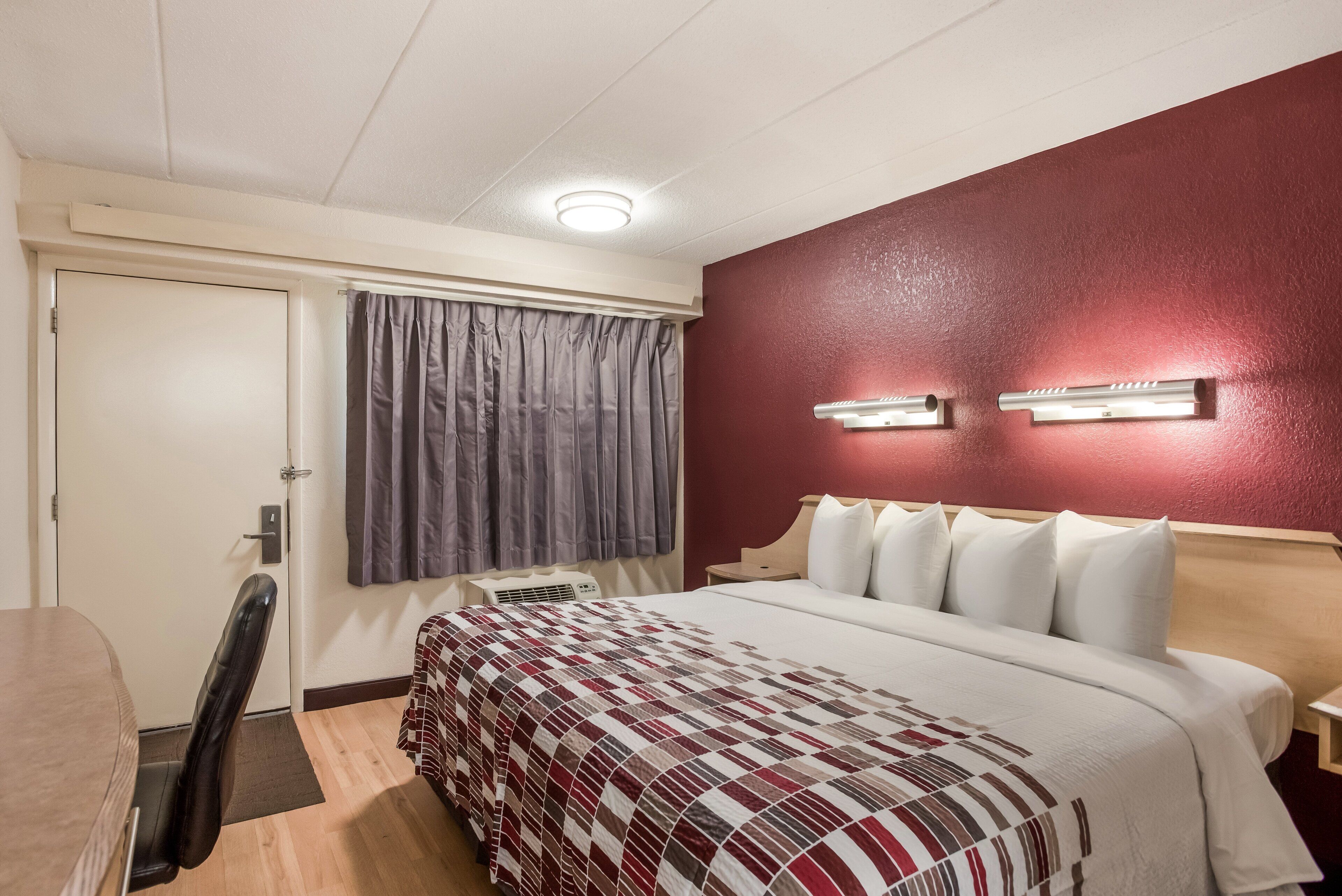 Habitación de hotel con cama, escritorio, silla, ventana con cortinas y un calentador fijado a la pared.