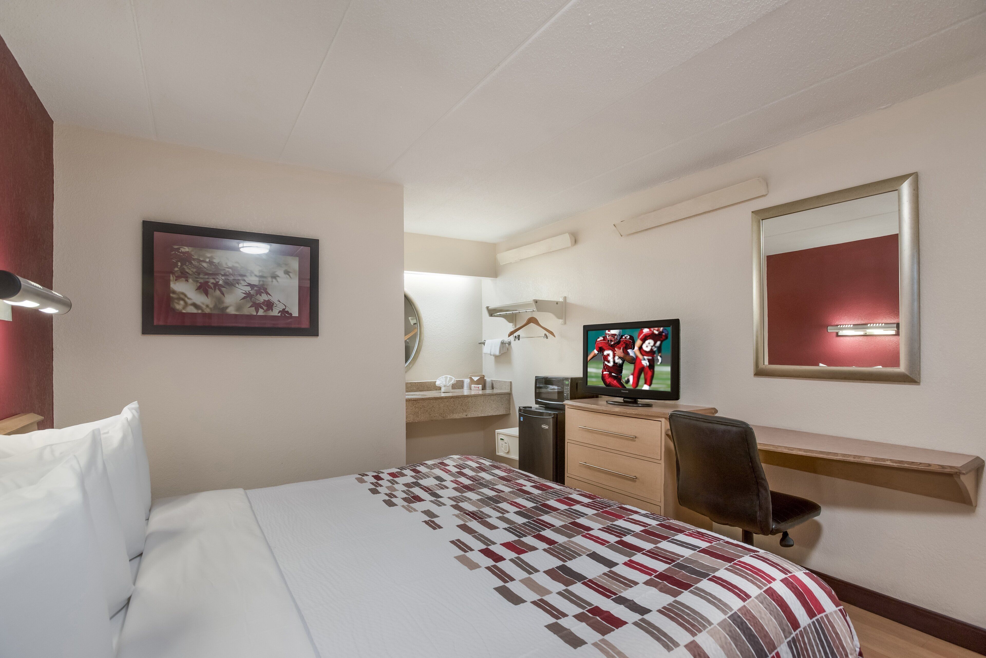 Ein Hotelzimmer mit einem Bett, einem Schreibtisch, einem Sessel, einem Fernseher, einem Spiegel und einem gerahmten Bild an der Wand.