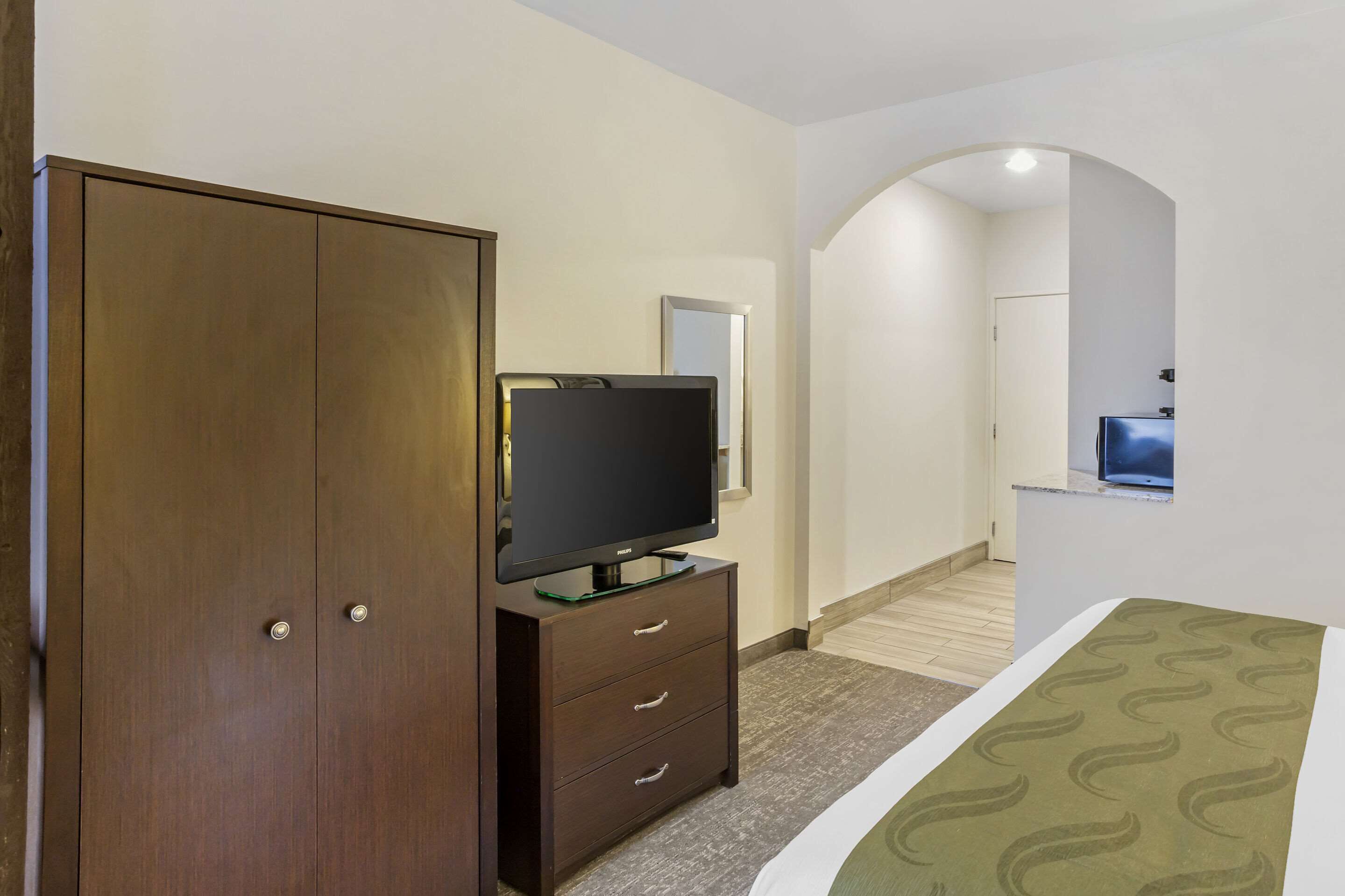 Une chambre d’hôtel avec une armoire en bois, un téléviseur posé sur un meuble, un lit recouvert d’une couvre-lit à motifs et un miroir au mur.