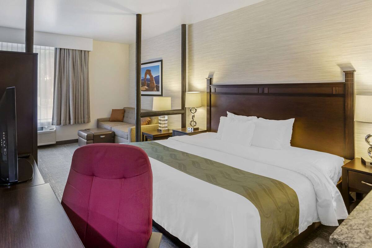 Una habitación de hotel con una cama grande, un sillón rojo, un escritorio con computadora, televisor y una ventana con cortinas.