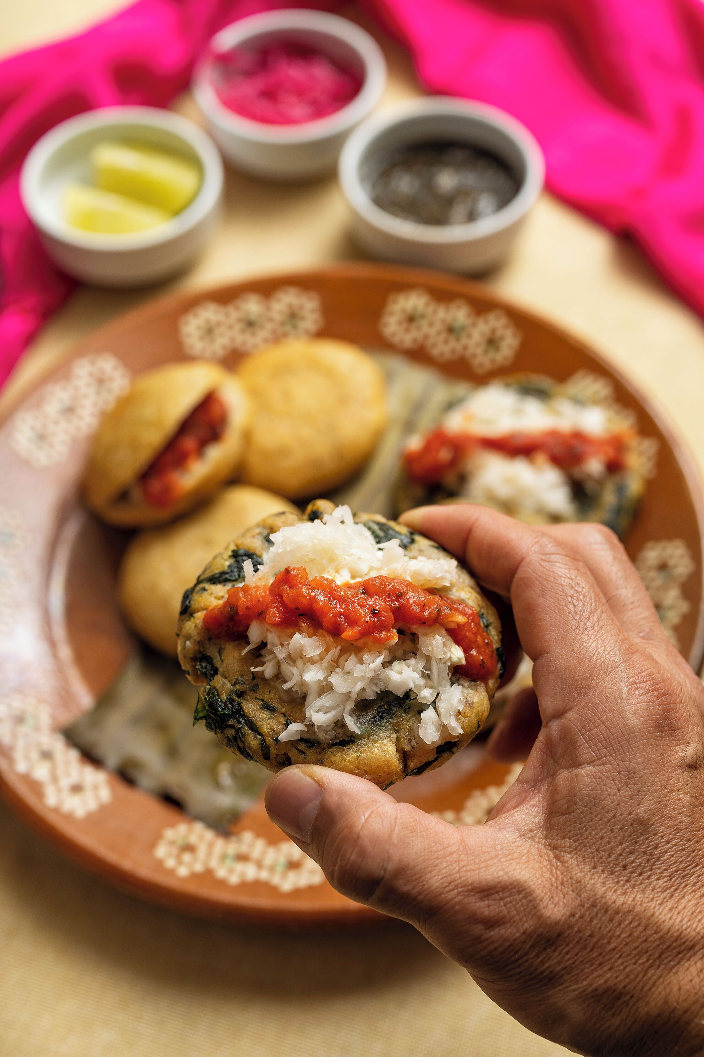 Una mano sostiene un pastel relleno de arroz y salsa de tomate, con otros pasteles y condimentos al fondo.