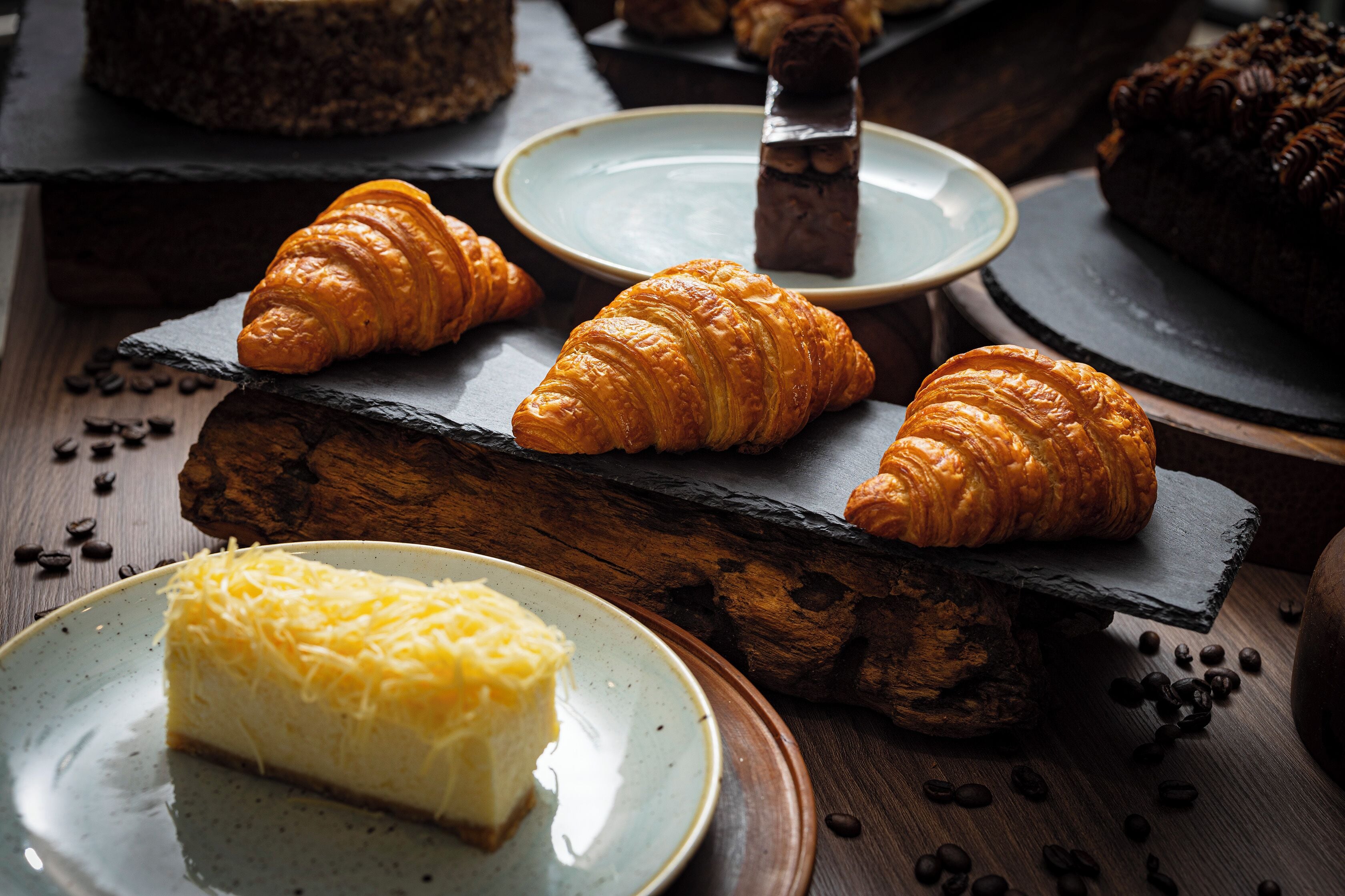 Variedad de pasteles en tablas de pizarra, incluyendo cruasanes y una porción de pastel de queso, con granos de café esparcidos alrededor.