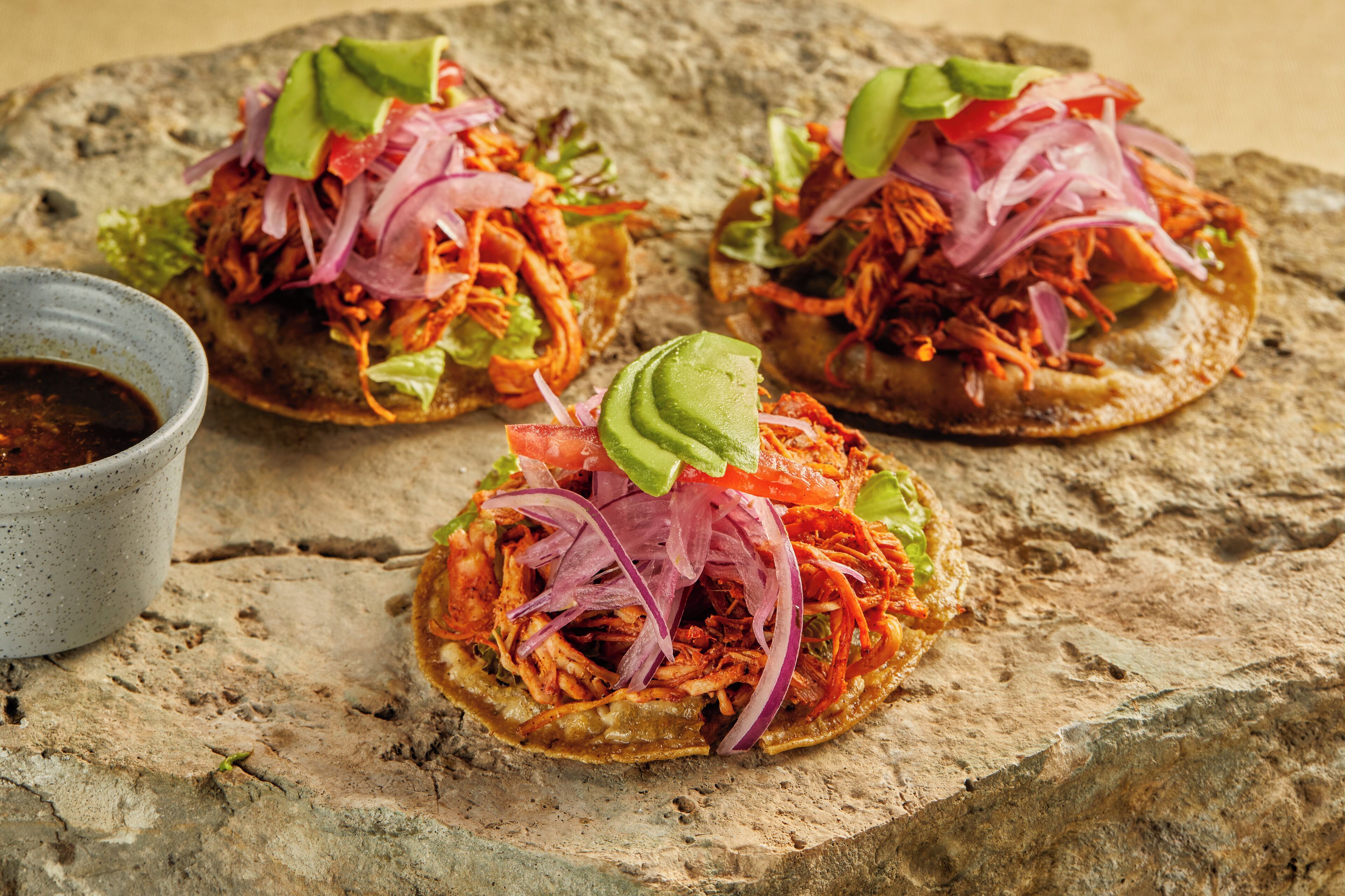 Tres tacos sobre una superficie de piedra con un tazón pequeño de salsa.