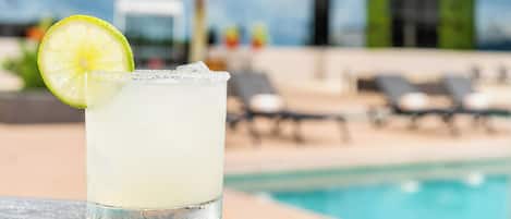 Un vaso de una bebida transparente con una rodaja de lima, sobre una mesa cerca de una piscina con sillones reclinables y palmeras al fondo.