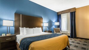 Quarto de hotel com cama grande, duas mesas de cabeceira, uma cadeira, janela com cortinas e luminária na parede.