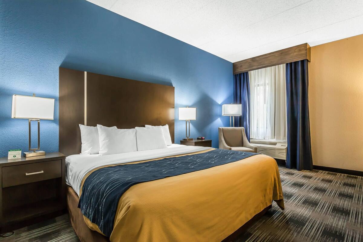 A hotel room with a large bed, two bedside tables, a chair, a window with curtains, and a wall-mounted lamp.