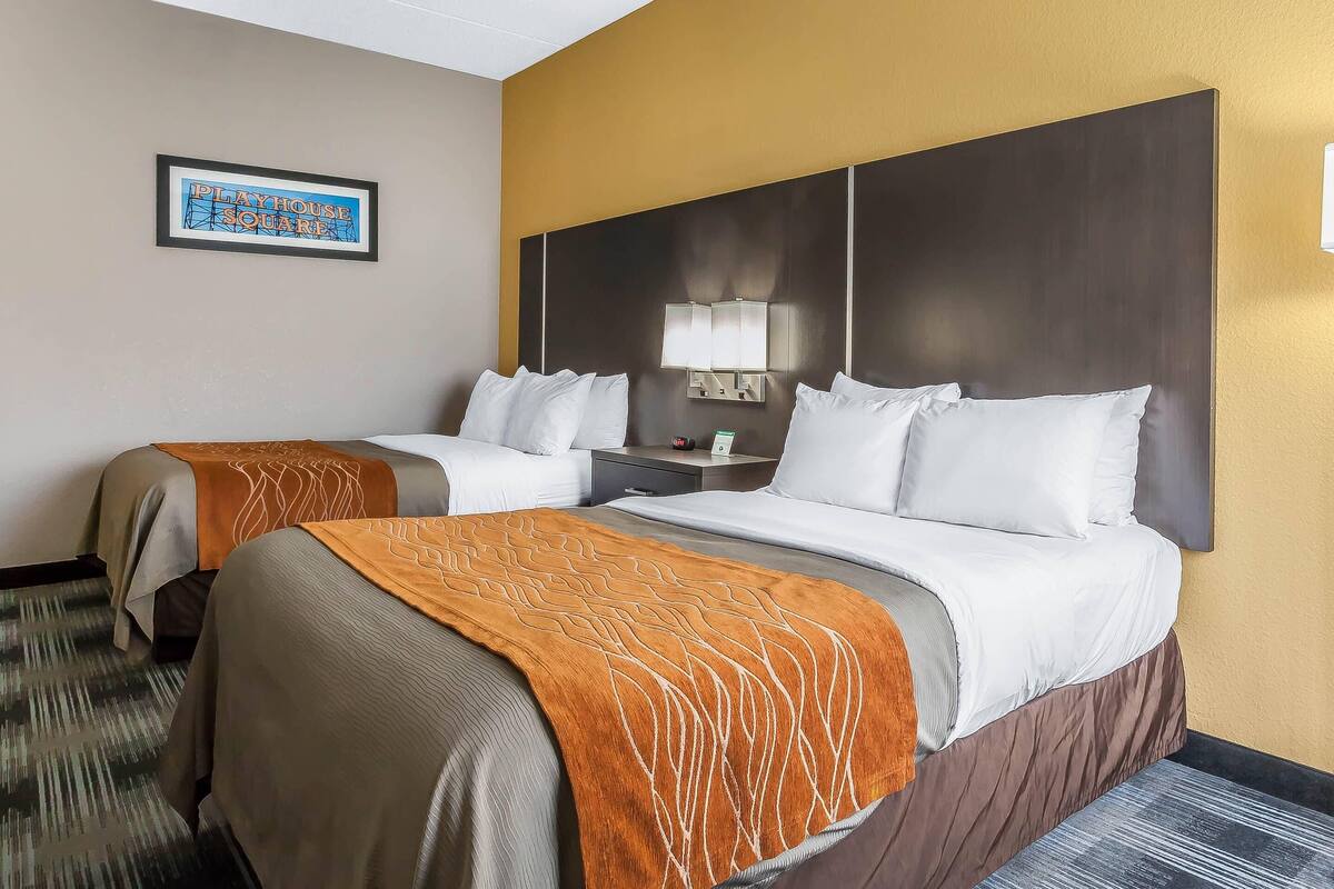 A hotel room with two beds, a headboard with a mirror, a framed picture on the wall, and a nightstand with a lamp.