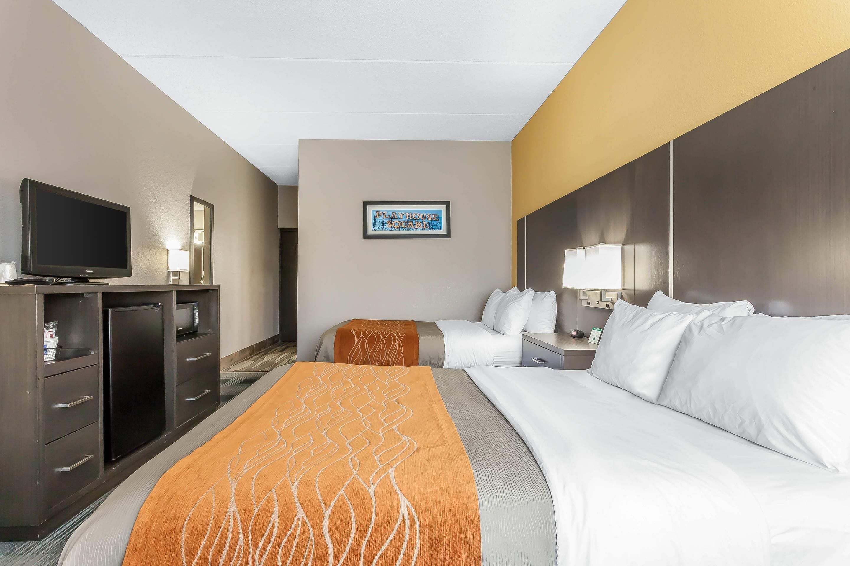 Quarto de hotel com cama, televisão, escrivaninha e mesa de cabeceira.