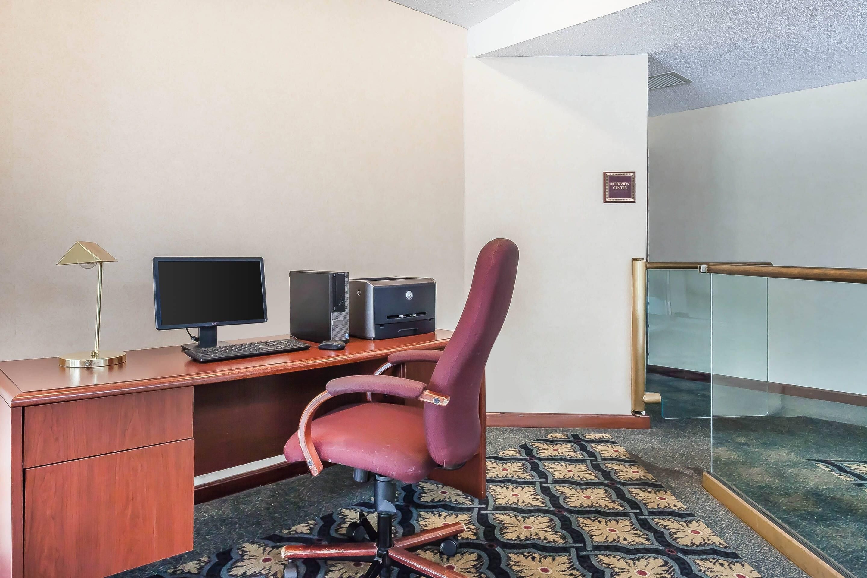 A hotel room with a wooden desk, a computer, a chair, a patterned carpet, and a glass partition.