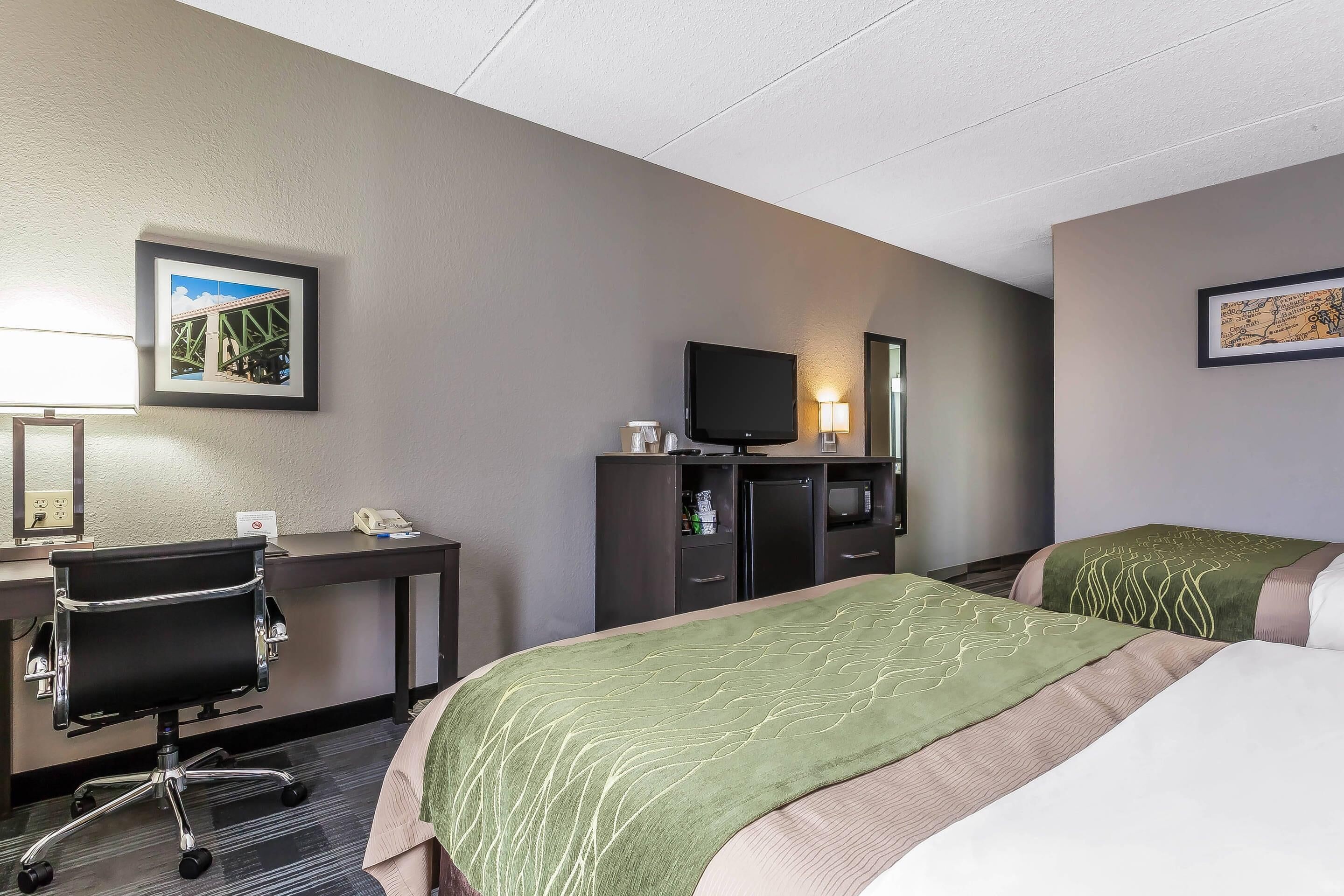 Una habitación de hotel con escritorio, televisor, dos camas con ropa de cama verde y beige, y cuadros en las paredes.