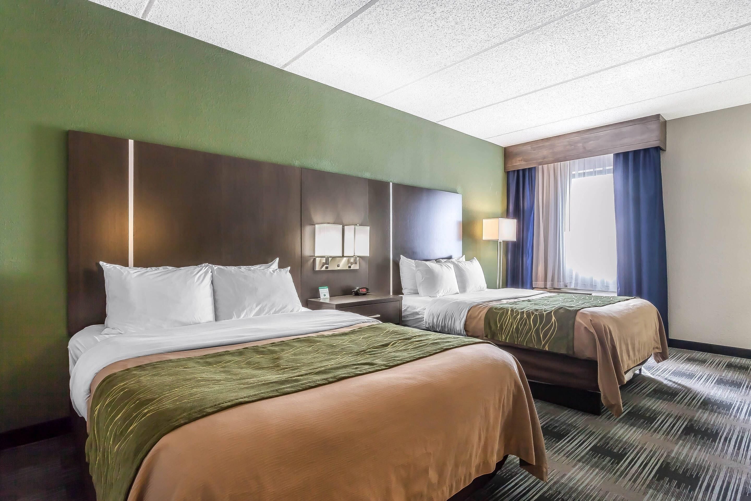 Quarto de hotel com duas camas, mesa de cabeceira com abajur e janela com cortinas.