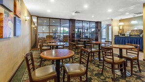 Um espaço de refeições de hotel com várias mesas e cadeiras, um buffet e grandes janelas que permitem a entrada de luz natural.