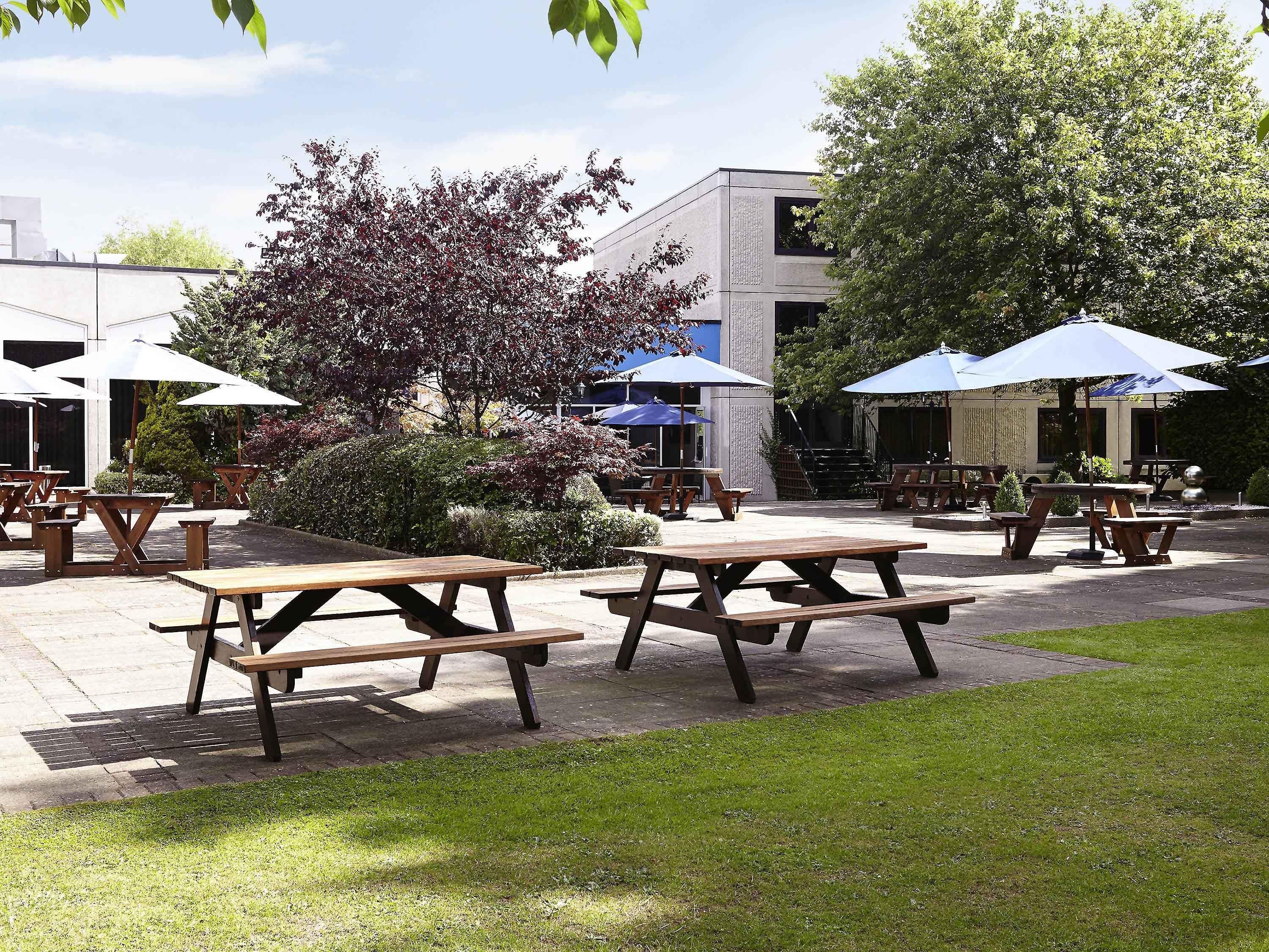 An outdoor patio area with picnic tables and umbrellas, surrounded by greenery and a building in the background.