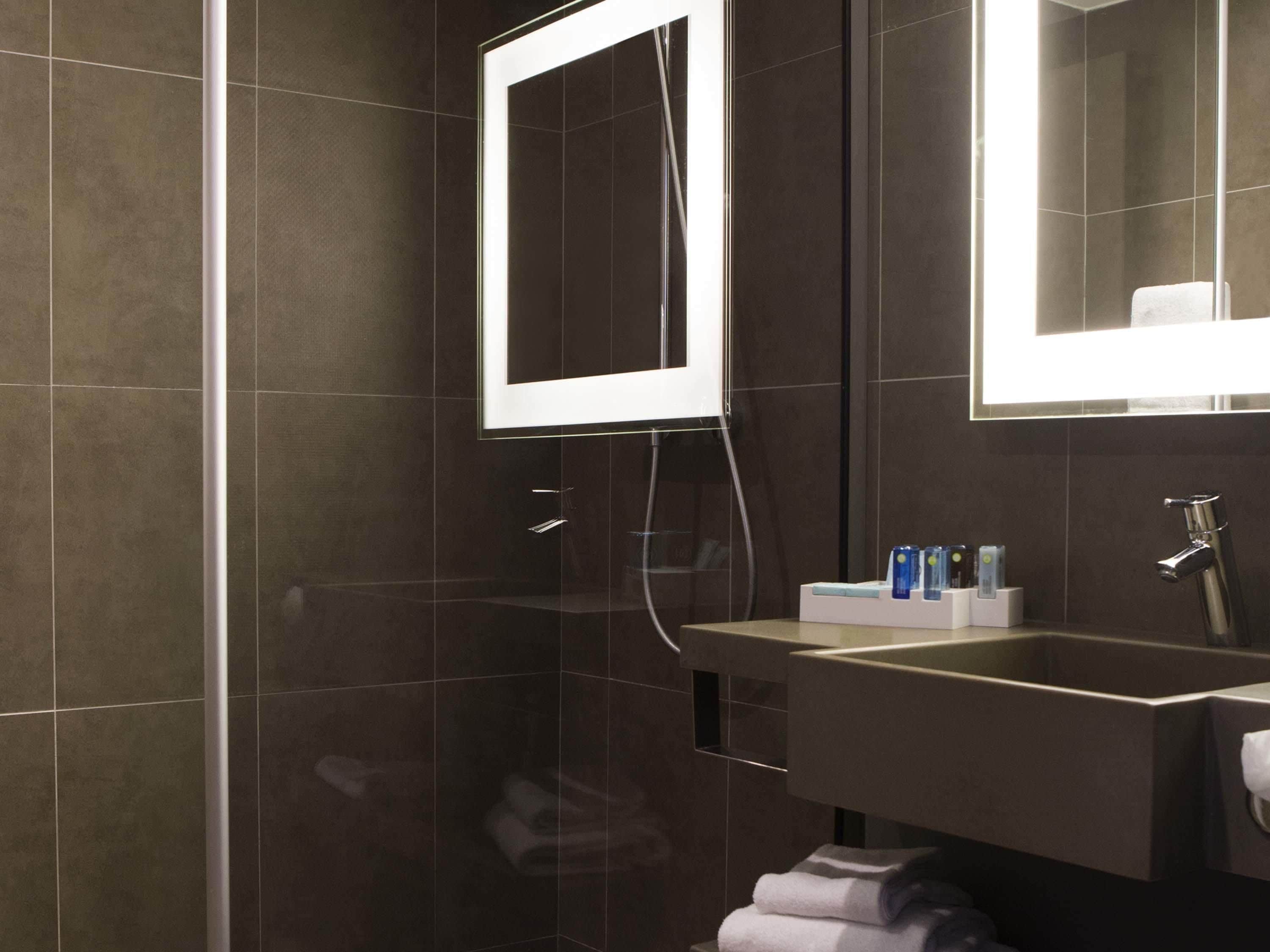 A modern bathroom with dark tiles, a glass shower enclosure, a sink with toiletries, and a mirror.