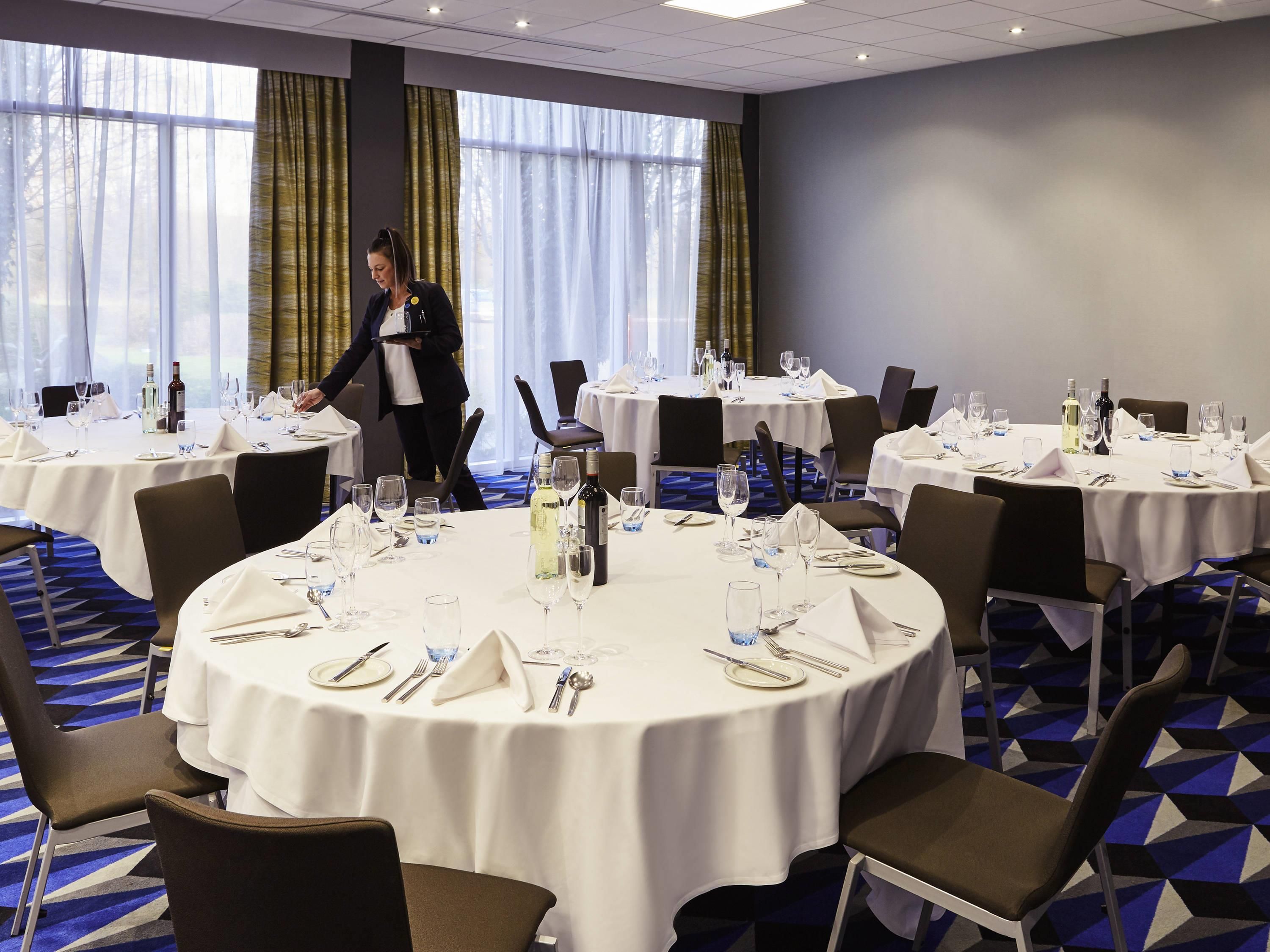A hotel dining area with round tables set for dining, a waiter in uniform, and large windows with curtains.