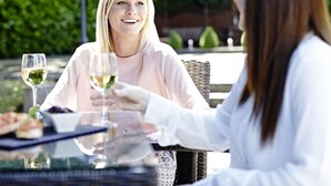 Two people seated at an outdoor table with a glass of white wine, a menu, and a wicker chair. - Novotel Coventry M6/J3 (Coventry)