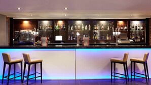 A bar area with a long counter, bar stools, and shelves stocked with bottles. - Novotel Coventry M6/J3 (Coventry)