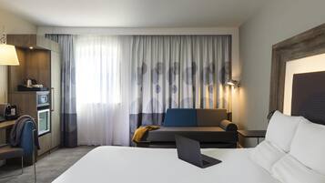 A hotel room with a bed, a desk with a laptop, a sofa, a coffee machine, and a TV.