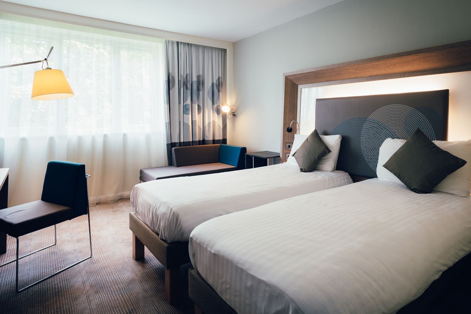 A hotel room with two beds, a desk, a chair, and a lamp.