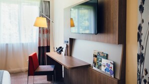 A hotel room with a bed, a desk with a chair, a lamp, a flat-screen TV mounted on the wall, and a window with curtains. - Novotel Coventry M6/J3 (Coventry)