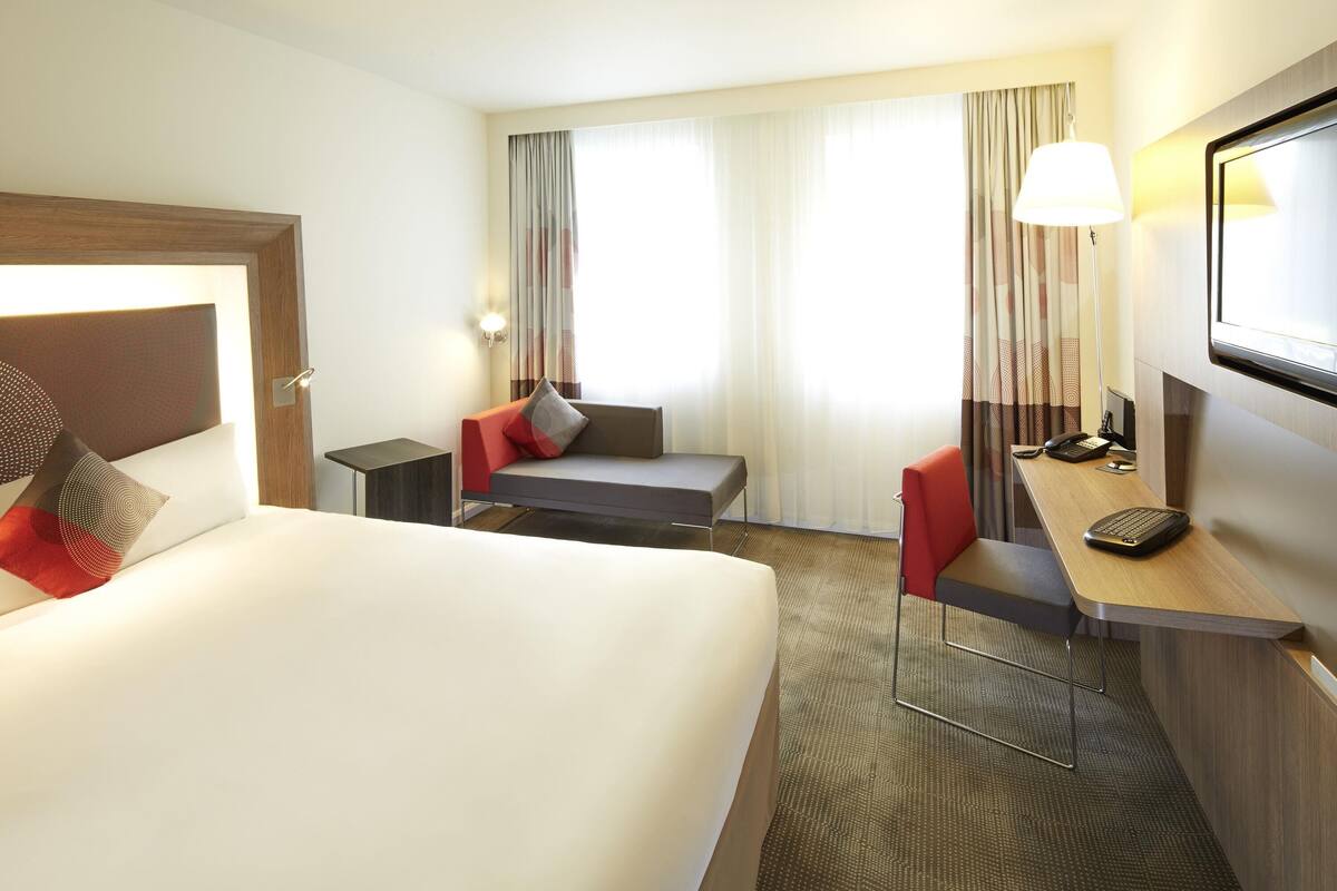 A hotel room with a bed, a desk, a chair, a lamp, and a TV.