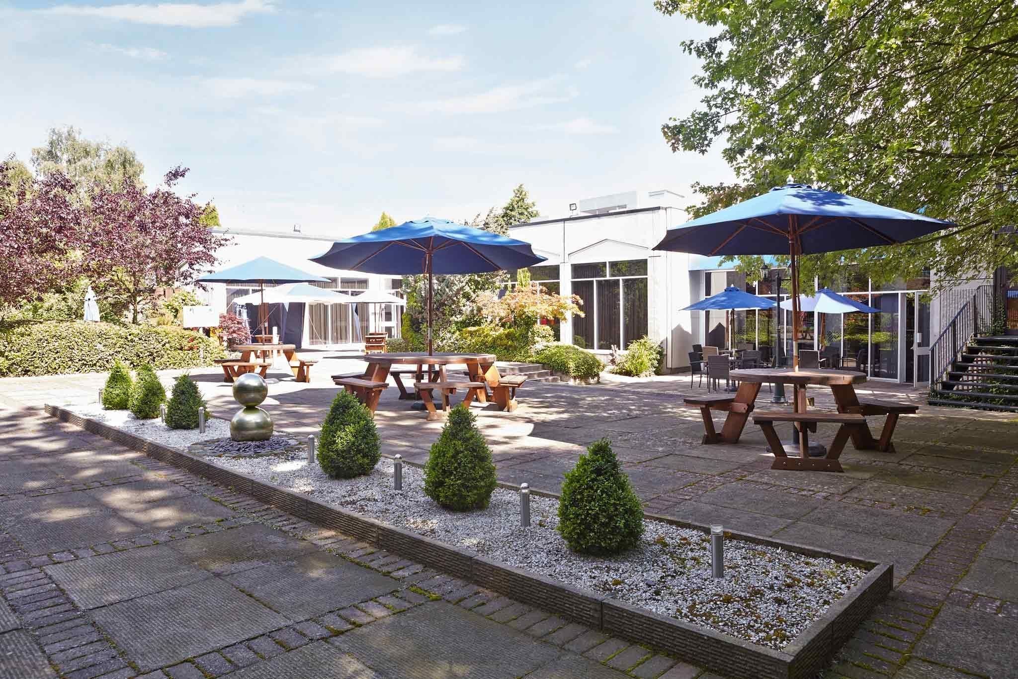 An outdoor patio area with picnic tables and blue umbrellas, surrounded by neatly trimmed hedges and a small garden.