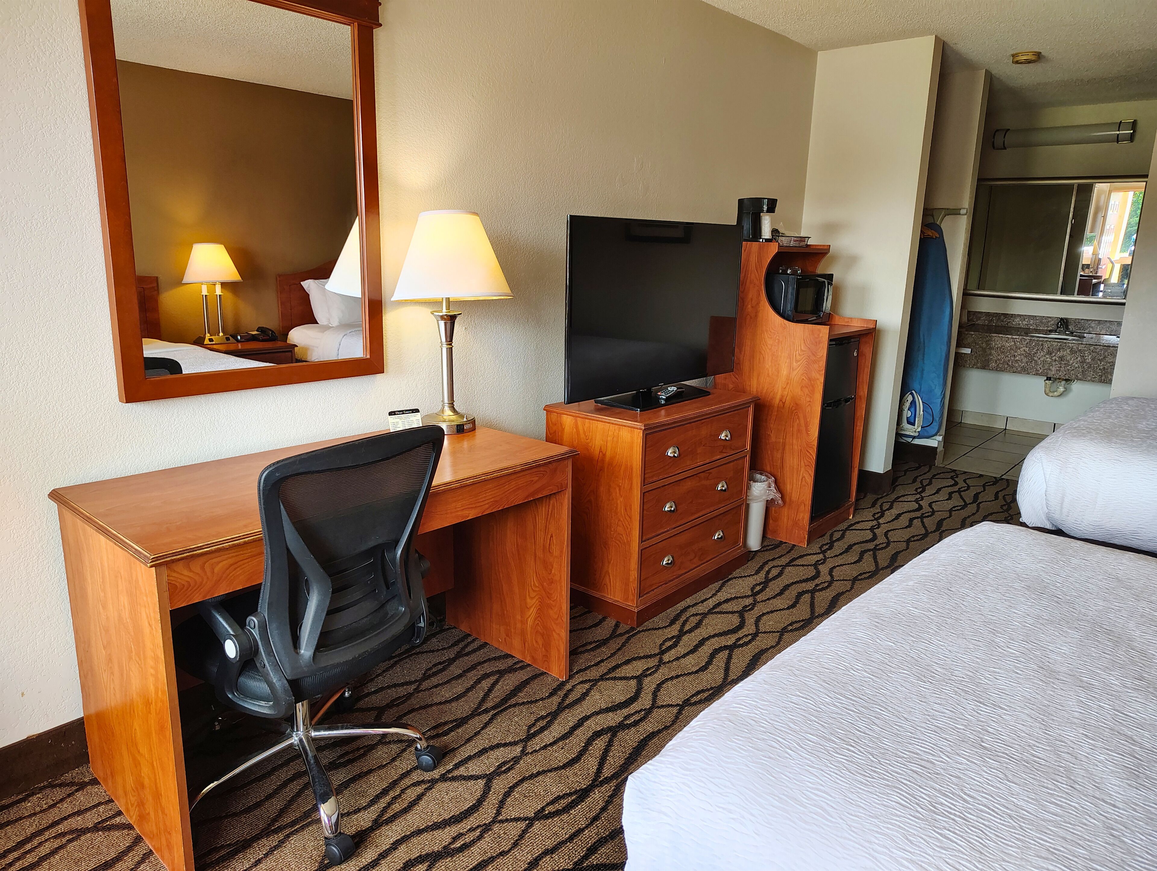 A hotel room with a wooden desk, a black office chair, a television, a minibar, and a bed.
