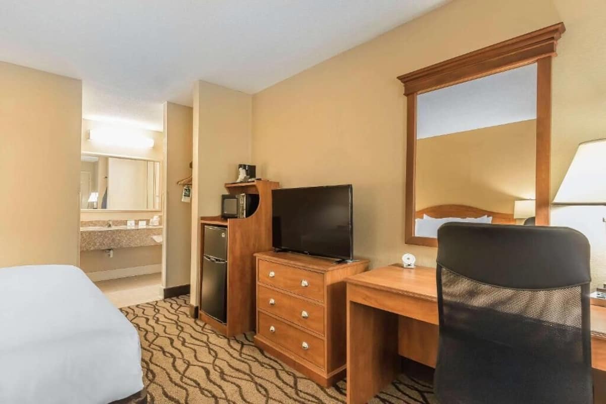 A hotel room with a bed, a desk with a computer, a TV, a dresser, a mirror, and a bathroom with a sink and mirror.