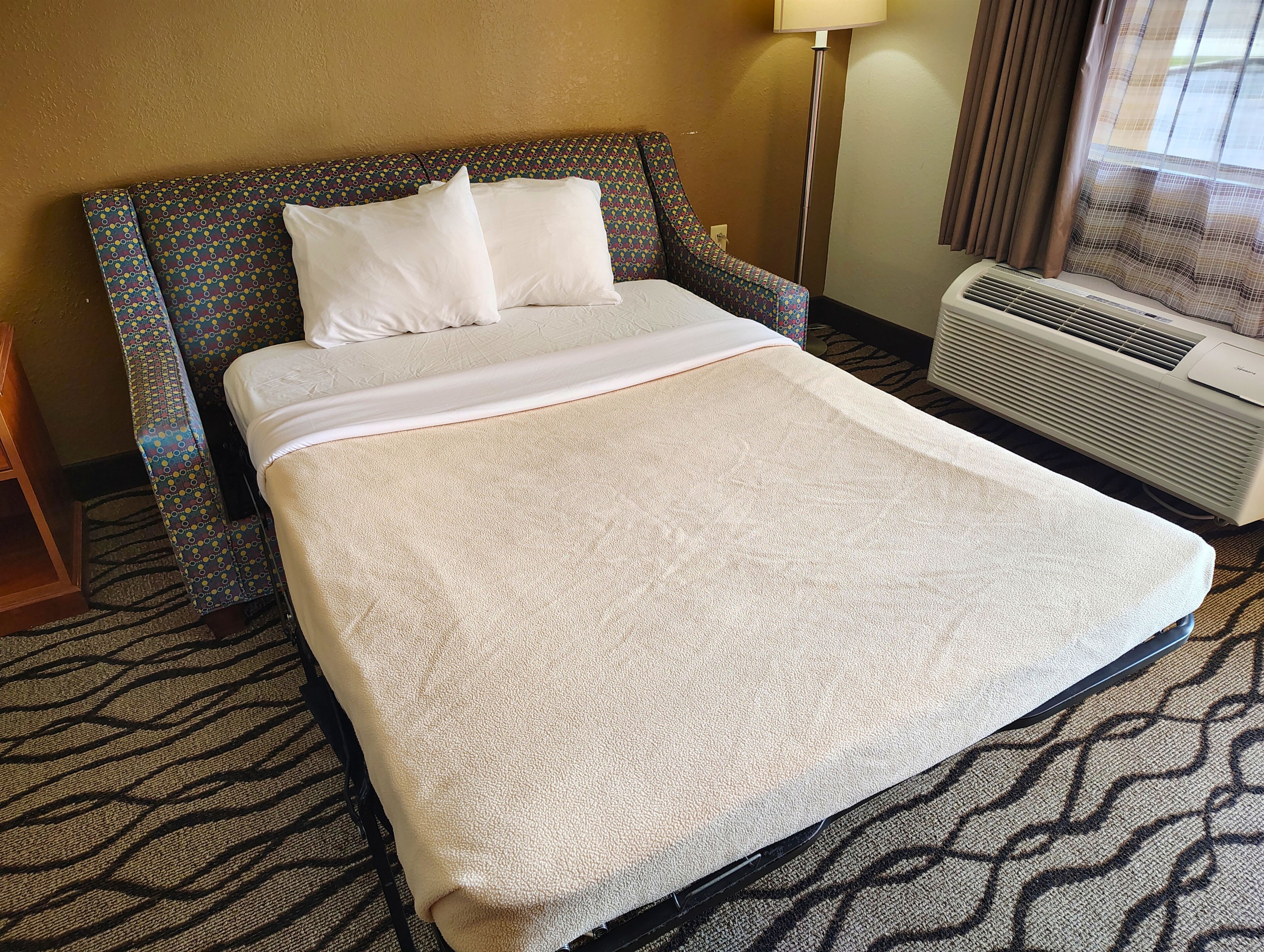A hotel room with a bed, a patterned armchair, a standing heater, and a window with curtains.