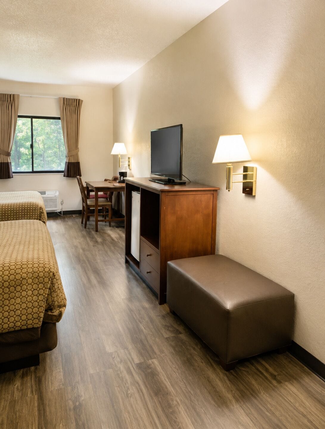 A hotel room with a bed, a desk with a television, a chair, a bench, and two wall-mounted lamps.