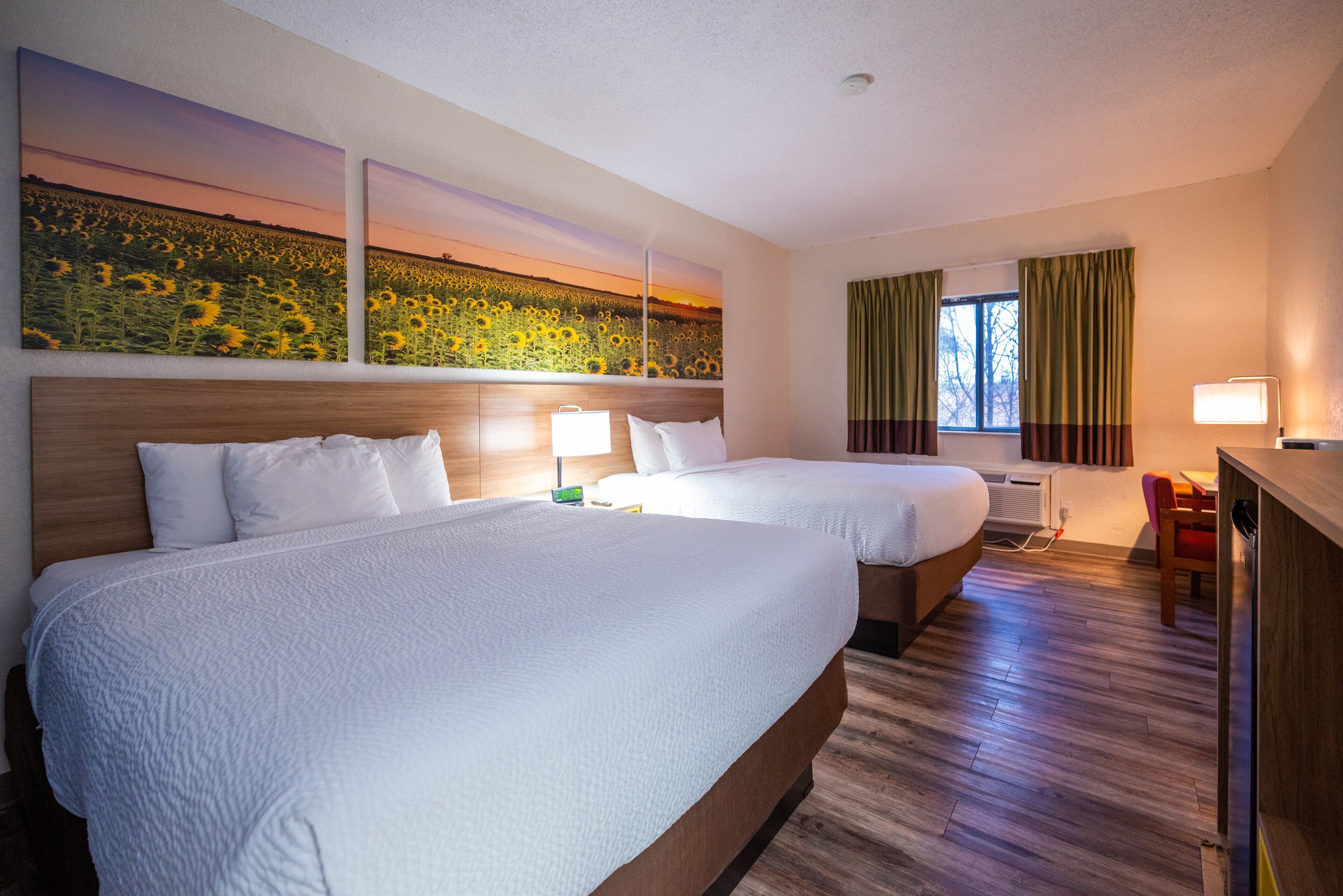 A hotel room with two beds, a desk, a chair, a window with curtains, and wall art.