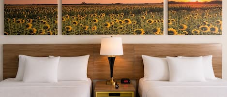A hotel room with two beds, a nightstand with a lamp, and a wall with three sunflower paintings.