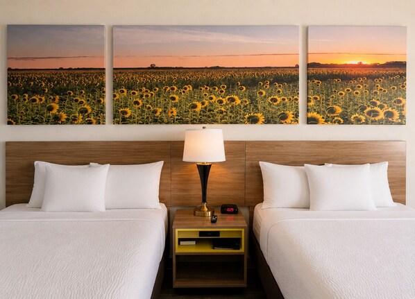 A hotel room with two beds, a nightstand with a lamp, and a wall with three sunflower paintings.