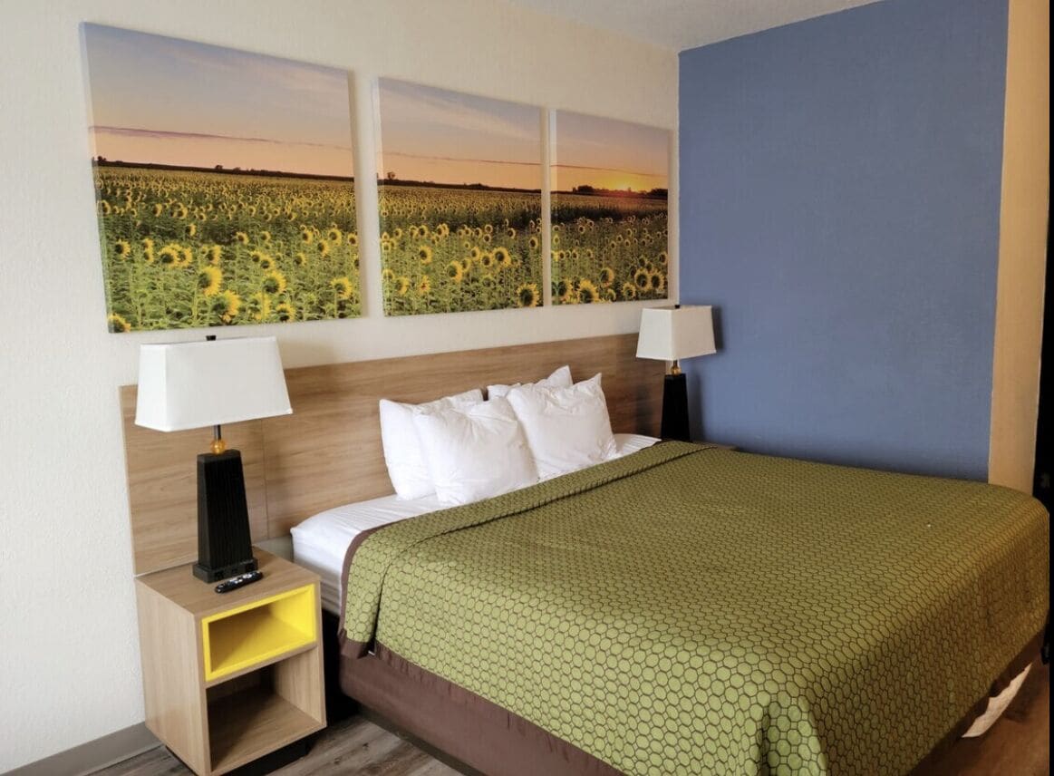 A hotel room with a bed, two bedside tables, a green bedspread, and three wall-mounted paintings of a sunflower field.