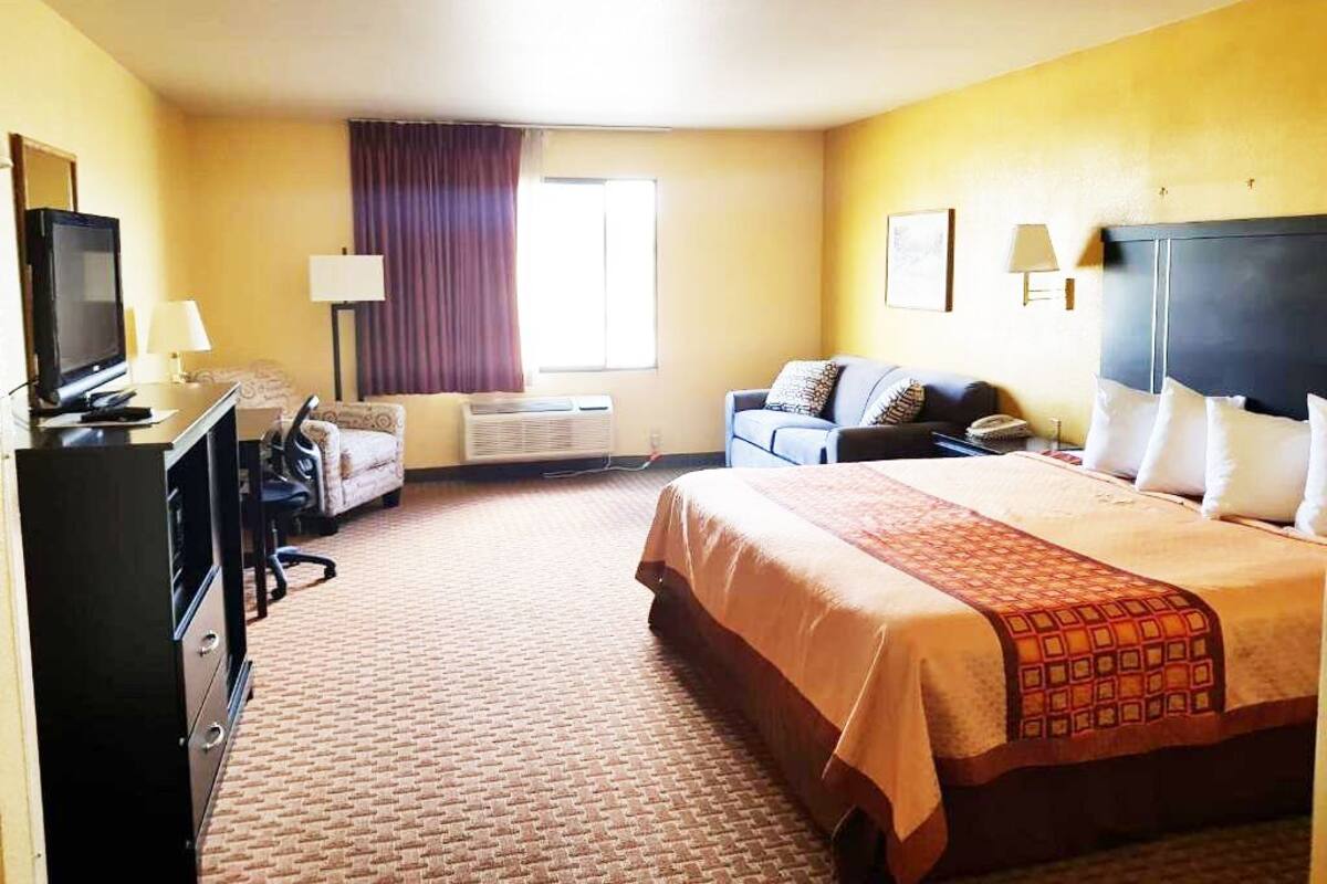 A hotel room with a bed, a desk with a computer, a television, a sofa, a window with curtains, and a wall-mounted lamp.