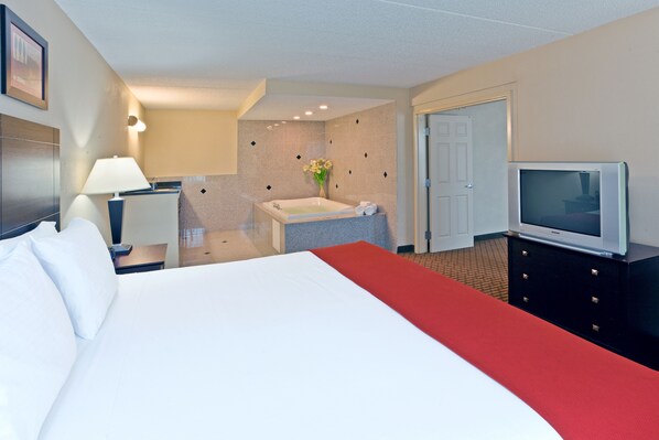 A hotel room with a bed, a TV, a desk with a lamp, a bathroom with a bathtub, and a door.
