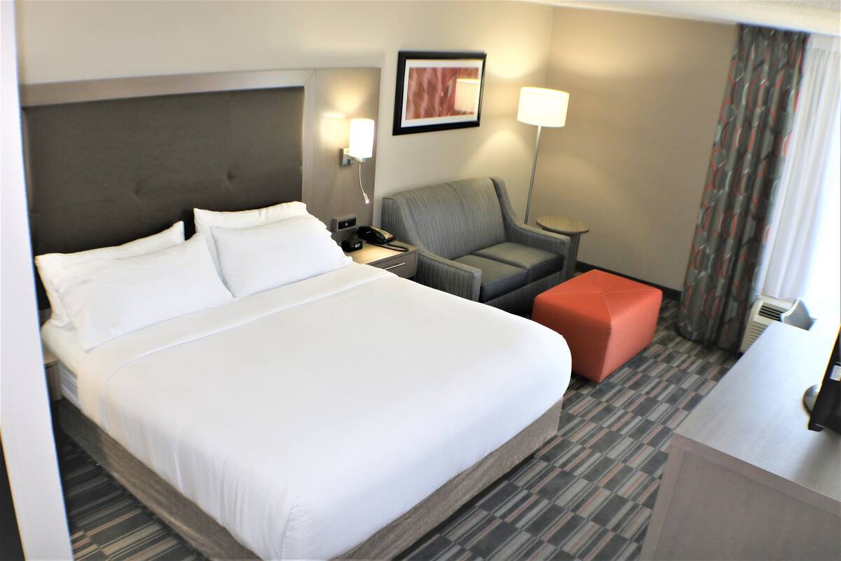 Ein Hotelzimmer mit einem großen Bett, einem grauen Sessel, einem kleinen roten Hocker, einem Schreibtisch und einer Lampe.