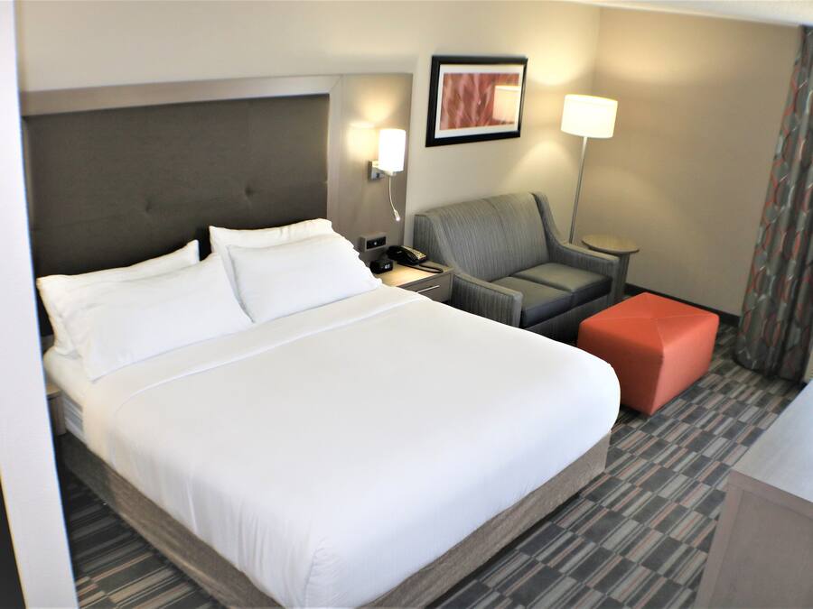 Habitación de hotel con una cama grande, un sillón gris, un reposapiés rojo pequeño, un escritorio y una lámpara.