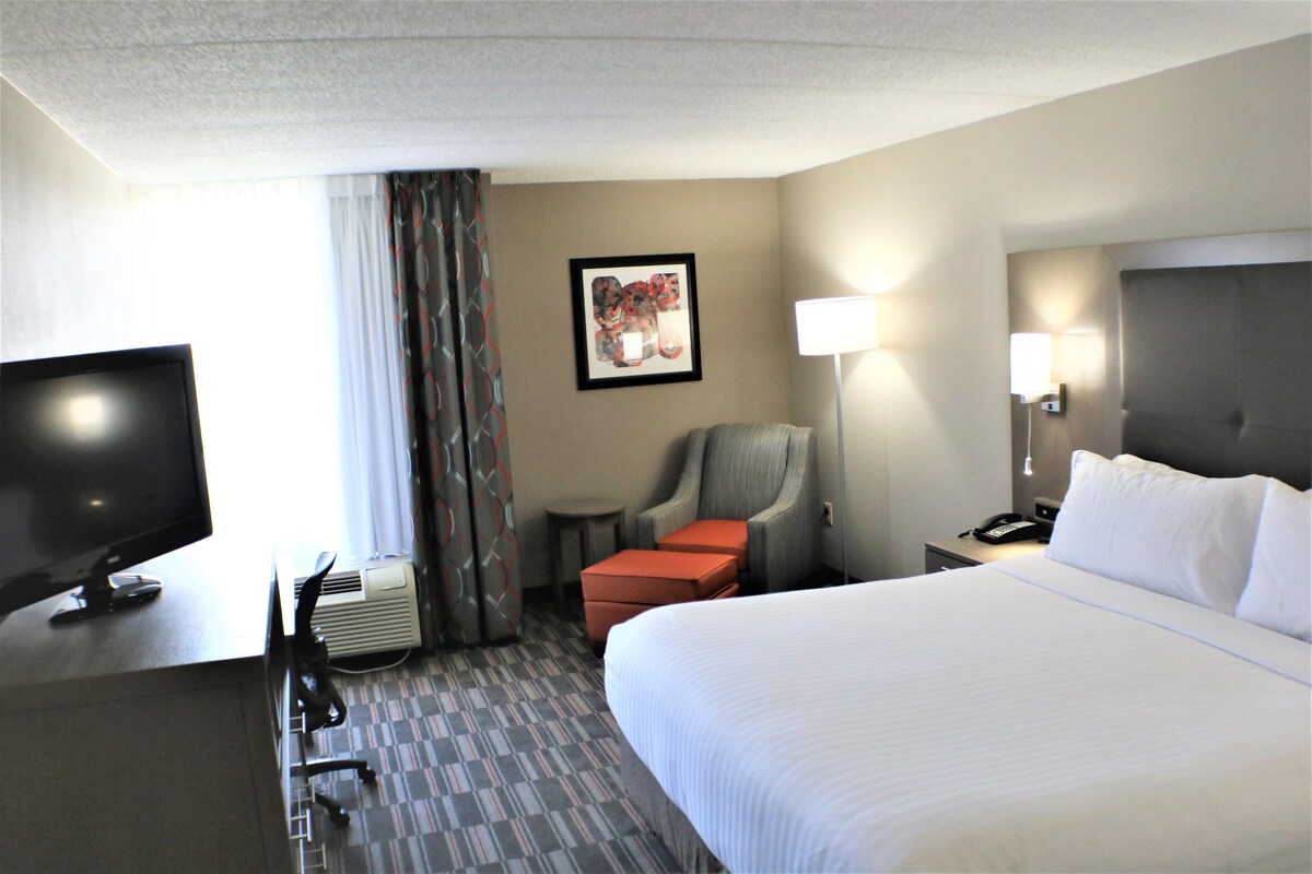 A hotel room with a large bed, a desk with a television, a chair, a small table, and a lamp.