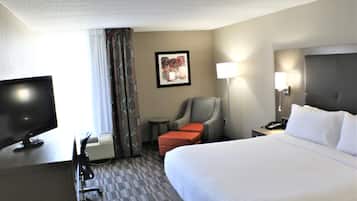 A hotel room with a large bed, a desk with a television, a chair, a small table, and a lamp.